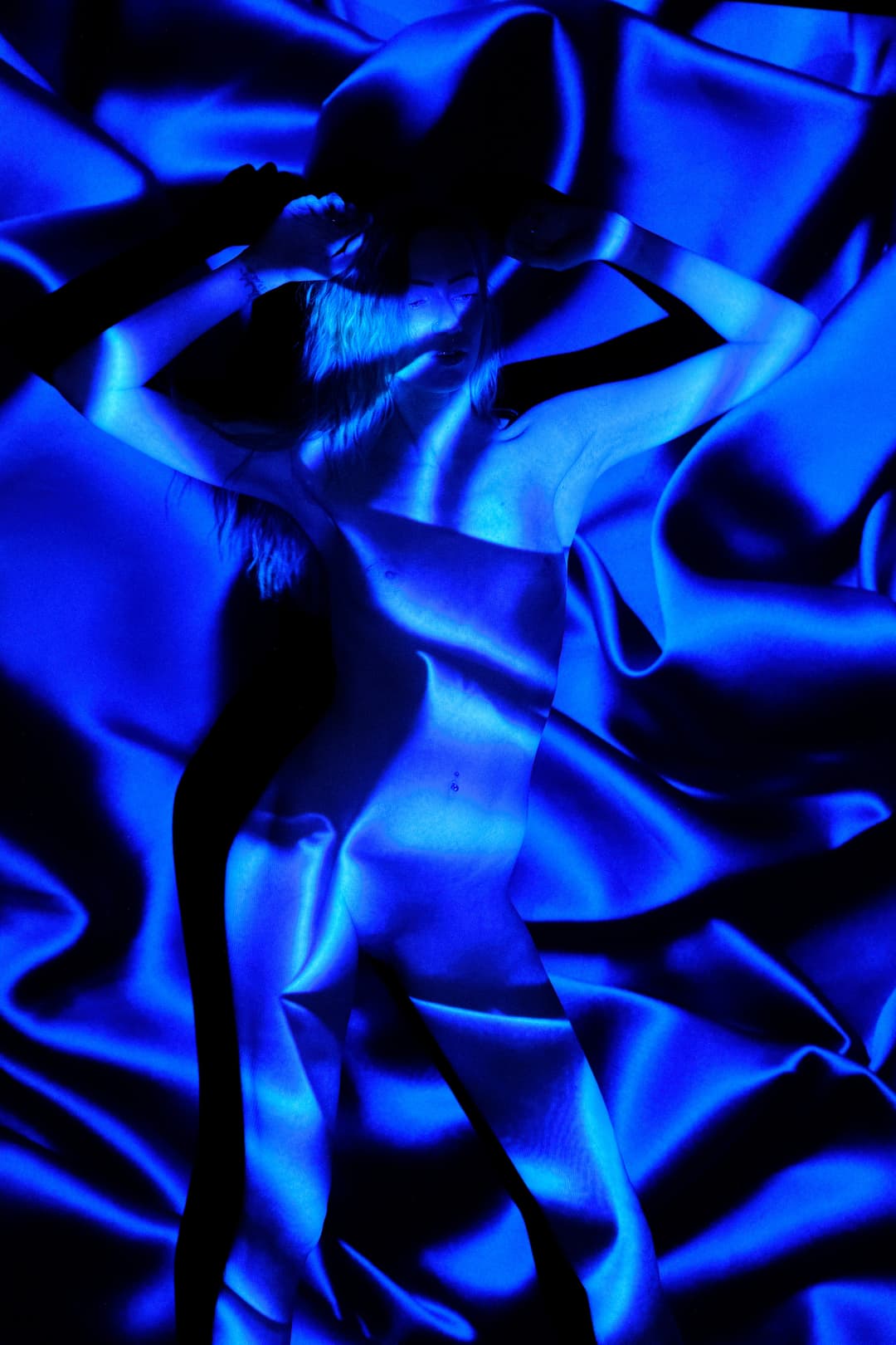 A person stands centrally against a background of deeply gathered, folds of royal blue silk fabric, bathed in intense blue light. Shadows cast from vertical slats or blinds create distinct, dark parallel lines across the person's upper body and face. The arms are raised with hands placed behind the head.