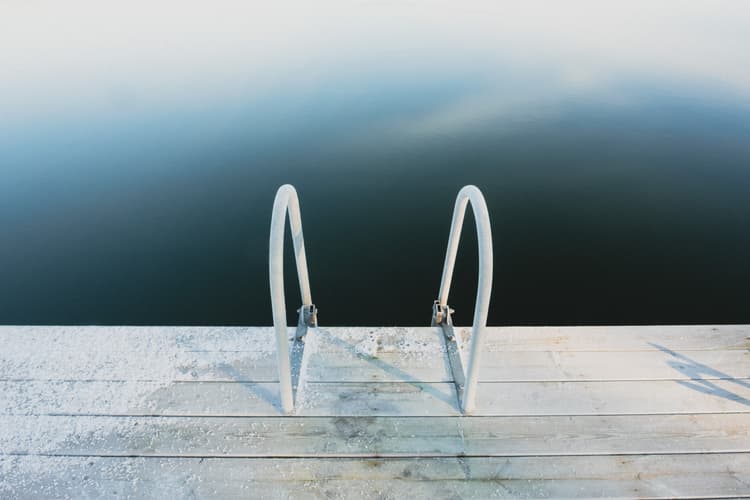 A wooden pier with a metal ladder extending down into dark, still water. A frost or thin layer of snow coats part of the wooden planks near the ladder, with soft light casting a long, sharp shadow of the ladder onto the deck.