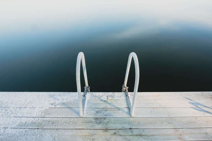 A wooden pier with a metal ladder extending down into dark, still water. A frost or thin layer of snow coats part of the wooden planks near the ladder, with soft light casting a long, sharp shadow of the ladder onto the deck.