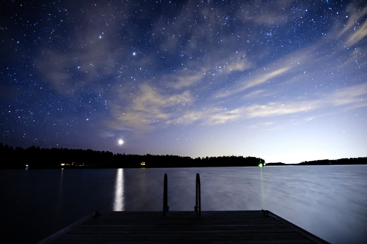 A night landscape view looking from a wooden dock out over a still body of water toward a distant dark treeline. A bright light from the sky, Venus, creates a sharp, vertical reflection on the dark water. The sky is filled with visible stars and wispy clouds, illuminated by the bright light.