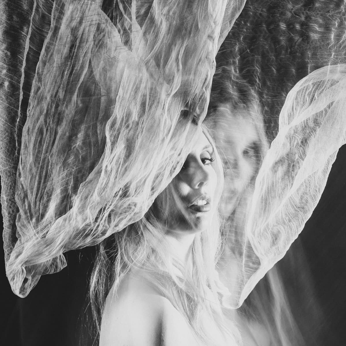 A black-and-white double exposure portrait showing a face partially obscured by flowing, semi-transparent fabric that drapes over the head and shoulders, creating a ghostly, layered effect in the composition.