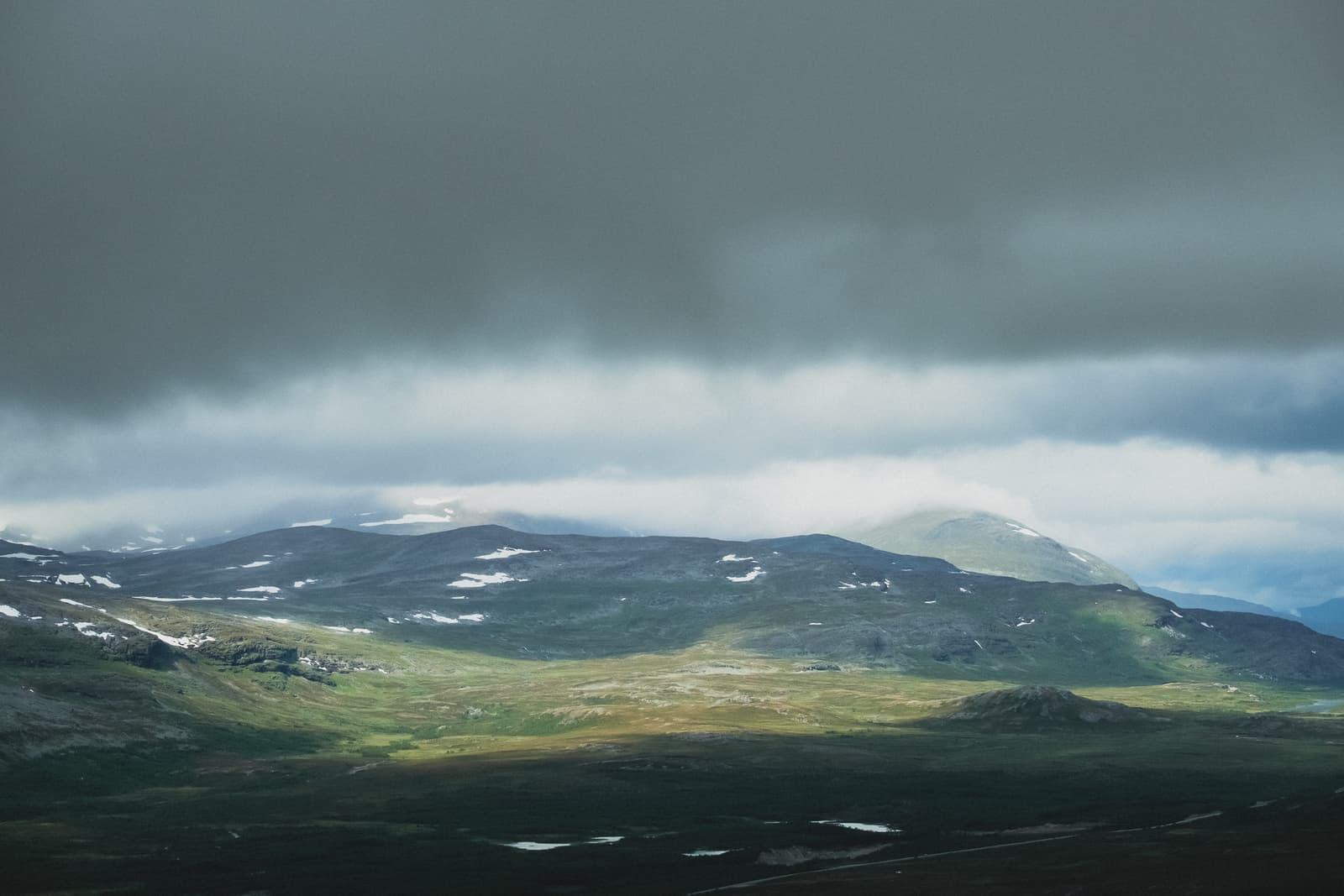 A wide, elevated landscape view in the Sami region under a heavy, low-hanging grey overcast sky. Patches of sunlight break through the clouds to illuminate sections of the tundra, which is marked by dark green vegetation, rocky ground, and small scattered patches of lingering snow on the rolling hills.