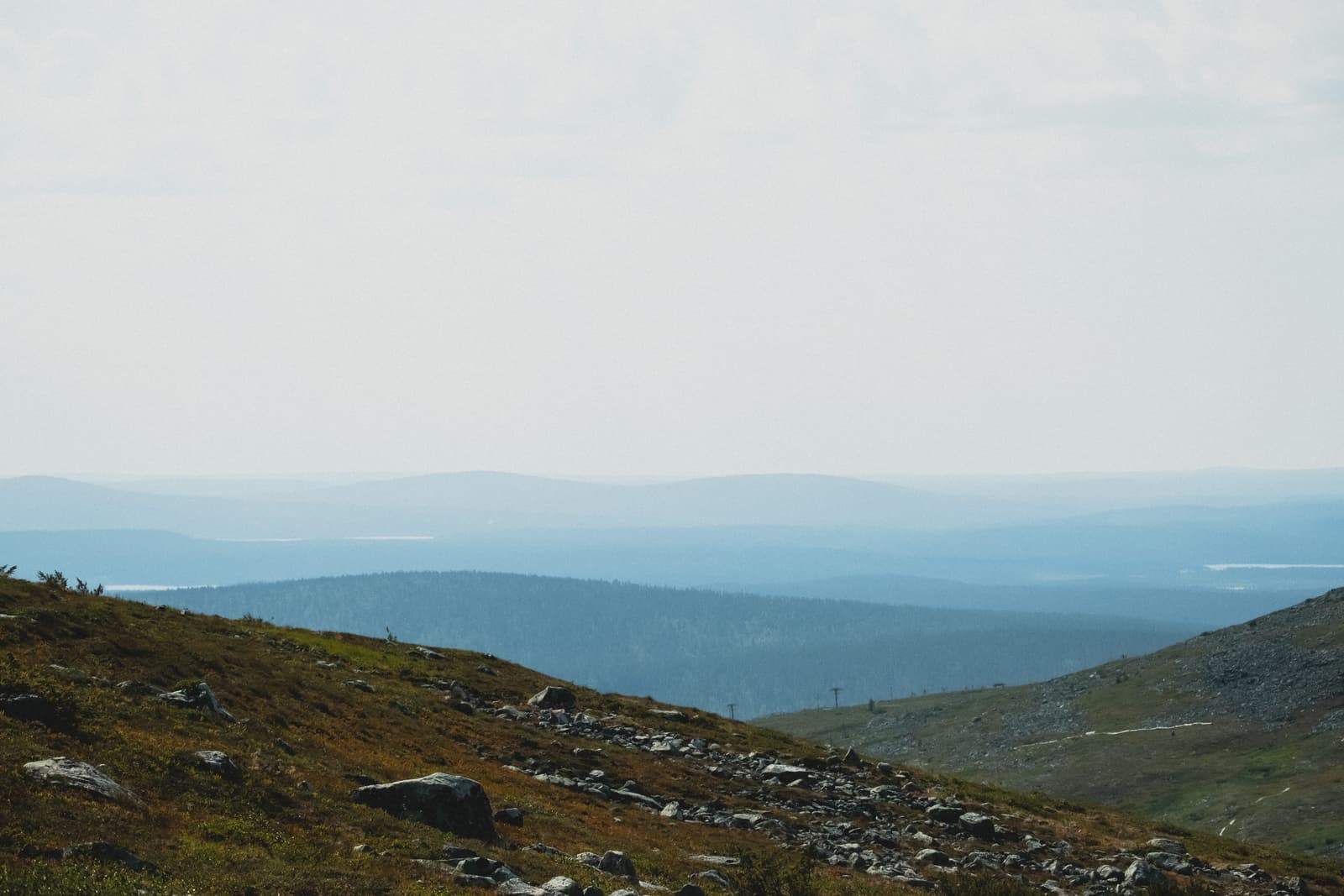 A wide, hazy landscape view of layered mountains and valleys in the Sami region. The foreground features a rocky, grass-covered hillside sloping downwards, while the distance shows multiple tiers of blue-toned mountains fading into a pale, overcast sky.