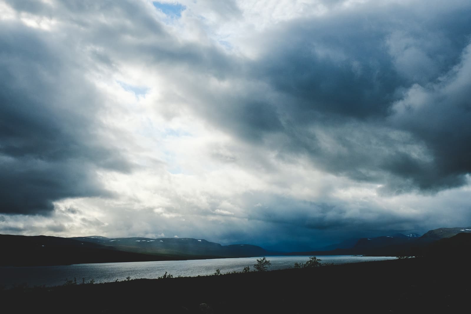 A wide landscape shot of a lake framed by distant, dark, rolling mountains under a heavy, stormy sky. Beams of light break through a gap in the dense gray clouds, illuminating a patch of the water's surface, while the foreground remains in shadow.
