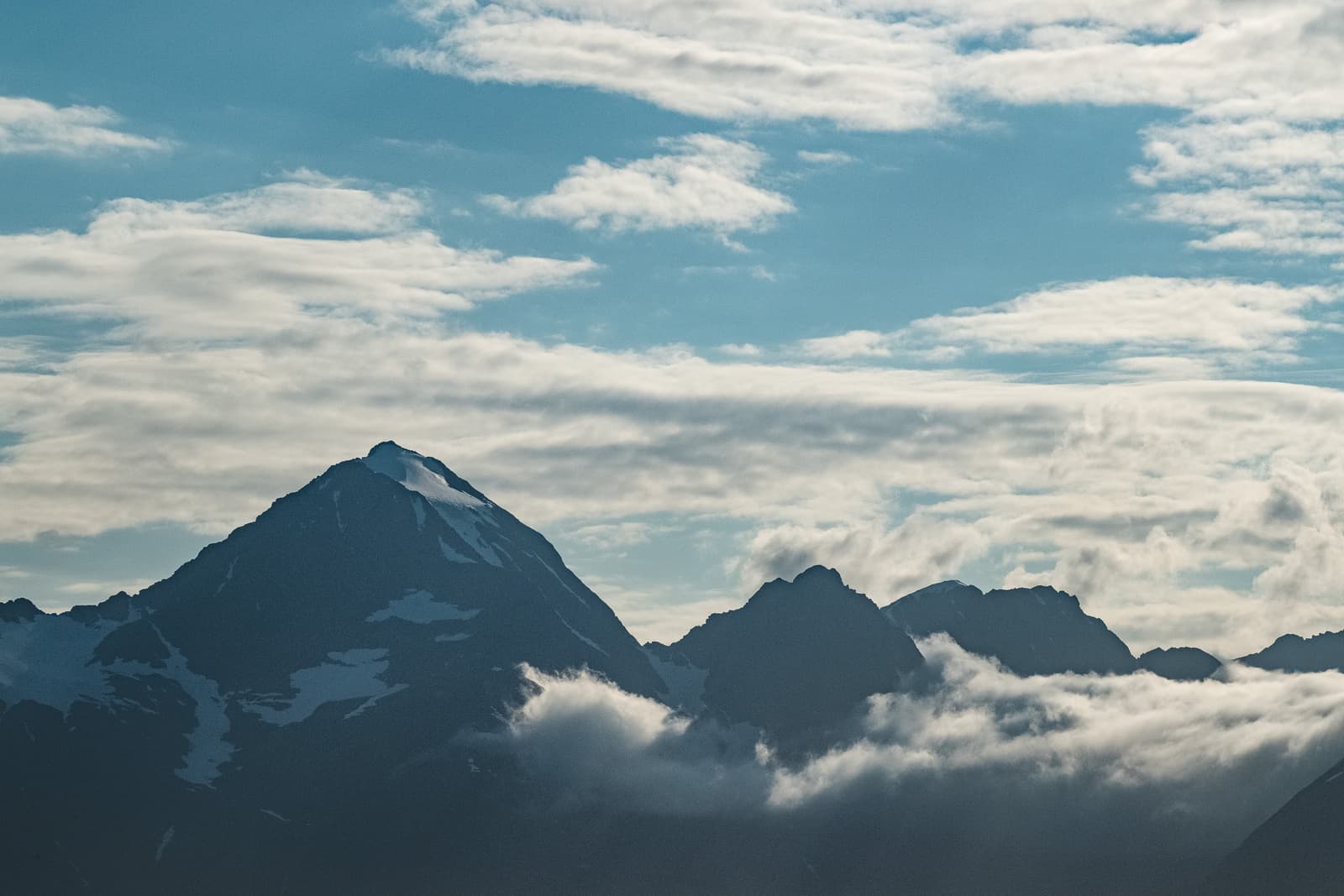 A wide shot of jagged mountain peaks under a bright blue sky filled with soft white clouds, with low-hanging fog clinging to the mountain bases and small patches of snow visible on the dark rock faces.