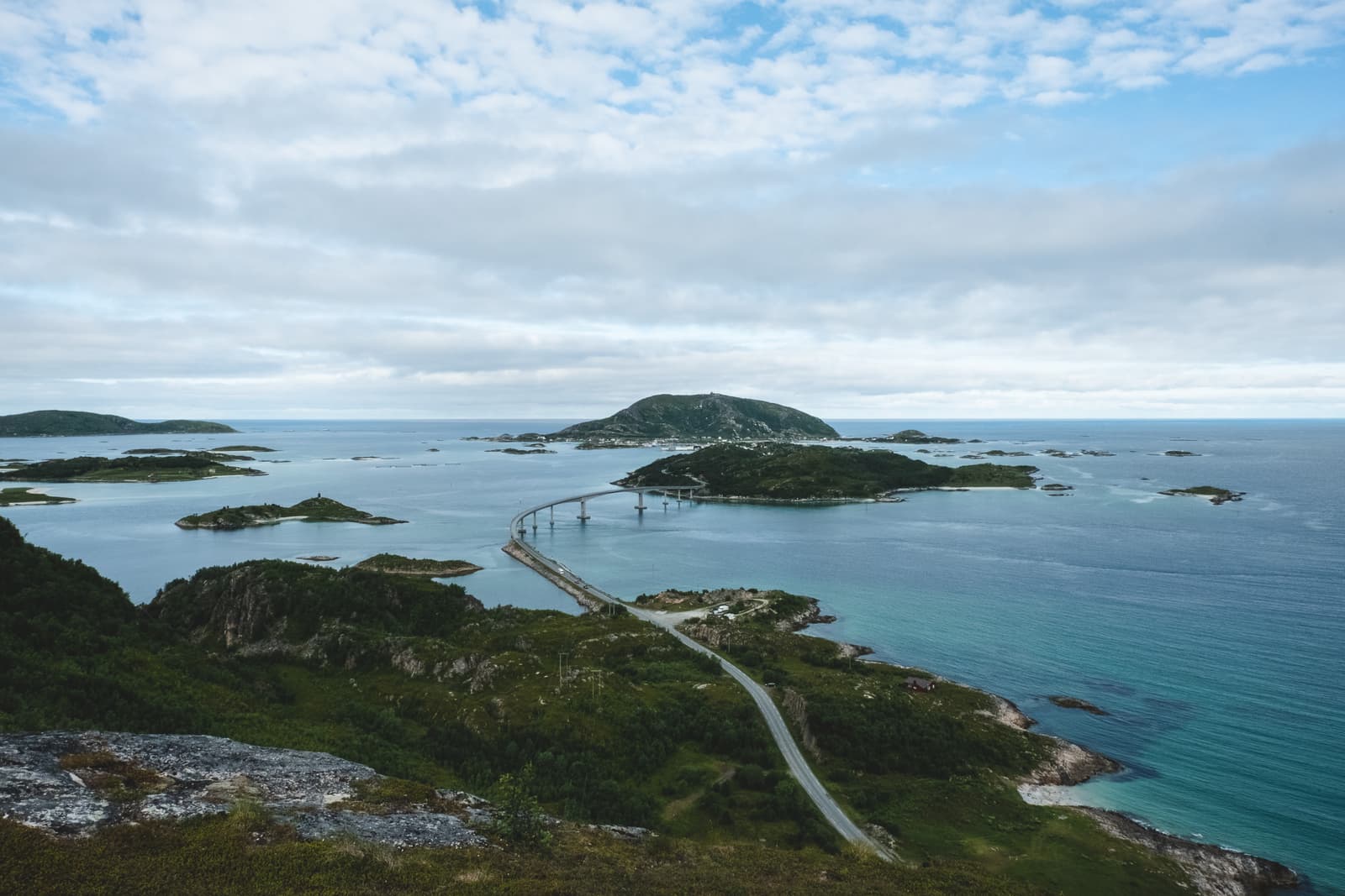 A high-angle landscape view of a coastal road in Northern Norway, featuring a bridge connecting small islands across a deep blue bay under a cloudy sky.
