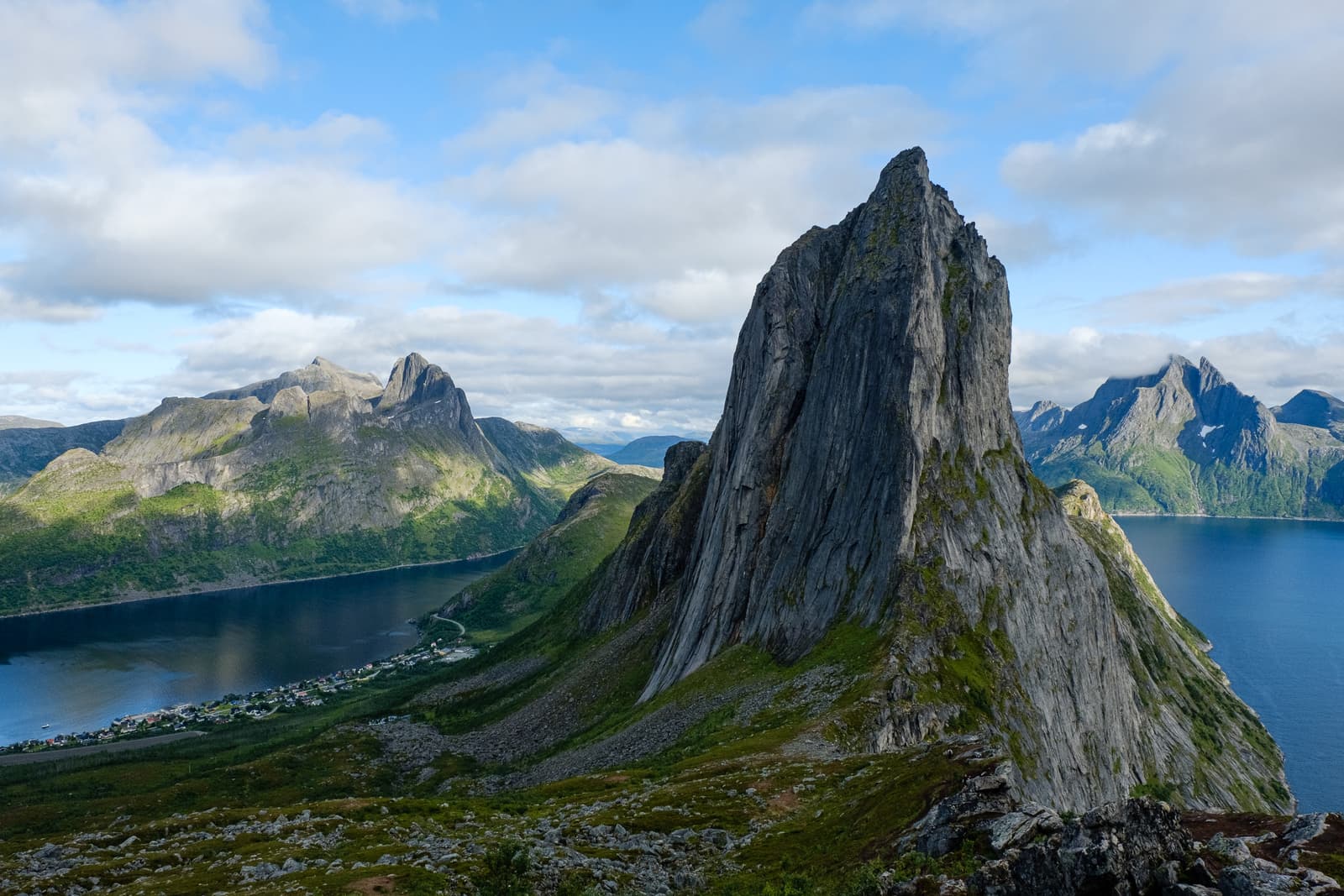 A wide, high-angle landscape shot of a towering, jagged granite peak rising above a deep blue fjord in Northern Norway. The foreground features mossy rock and low mountain scrub, leading down to a small town nestled along the water's edge. Beyond the central peak, more mountain ranges stretch into the distance under a blue sky with scattered white clouds.