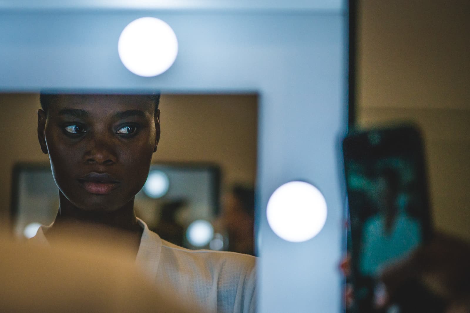 The reflection of a person in a vanity mirror, surrounded by bright circular bulbs. In the foreground, a handheld smartphone is visible in the frame, partially obscuring the view of the mirror. The lighting is dim, contrasted by the intense white light of the mirror bulbs.