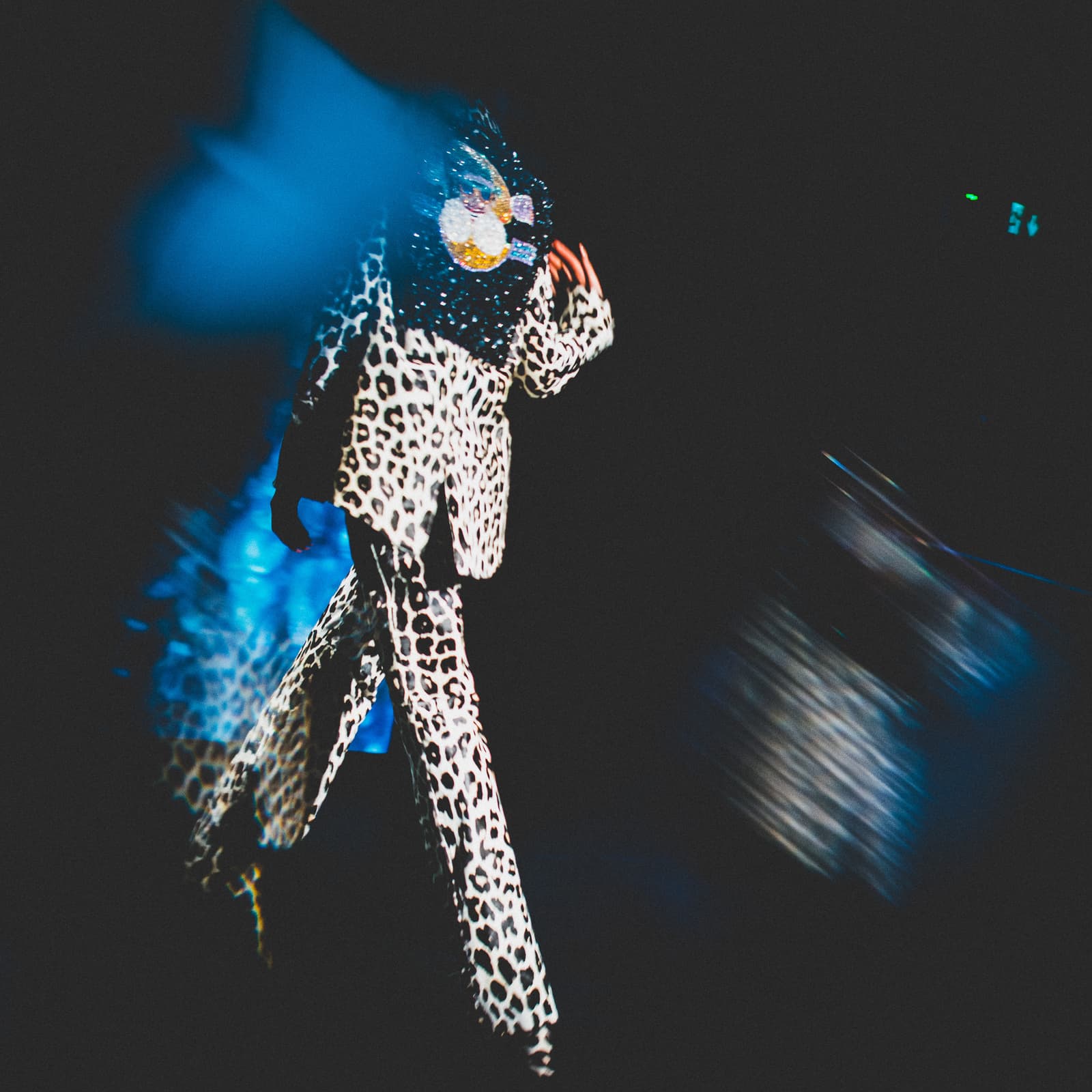 A long exposure photograph of a person walking on a runway wearing a leopard-print suit with a heavily beaded, dark collar. The motion blur creates streaks of blue light around the figure, against a solid black background.