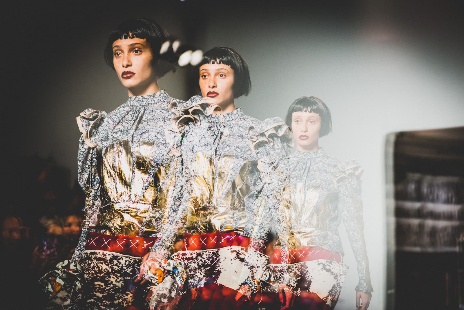 A triple-exposed image showing a model in a dark bob wig walking down a runway. The model wears a long-sleeved blouse with ruffled shoulders and a blue floral and gold metallic pattern, paired with a skirt featuring red, black, and white textures. The lighting is bright and directional, creating a glowing trail effect.