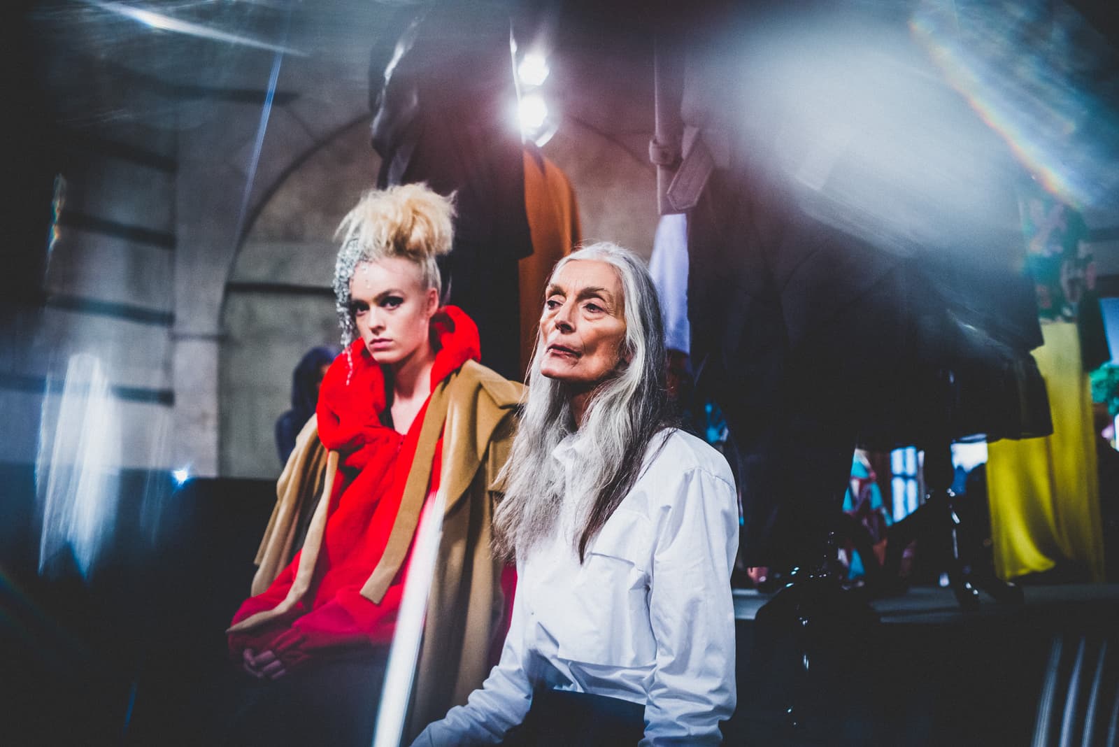 Two people are positioned in a backstage area filled with hanging garments. To the left, one wears a bright red hooded sweater under a tan trench coat, with blonde hair styled high. To the right, another person with long grey hair wears a crisp white collared shirt. Bright light flares across the top of the frame, obscuring details of the ceiling.
