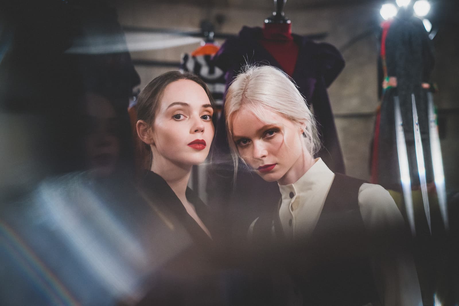A close-up shot of two individuals backstage at a fashion show, visible through a partially obscured foreground. One person has dark hair and wears a black garment; the other has platinum blonde hair and wears a cream-coloured high-neck shirt under a dark vest. Both wear bold red lipstick and gaze directly toward the camera, with garment racks and stage lights blurred in the background.