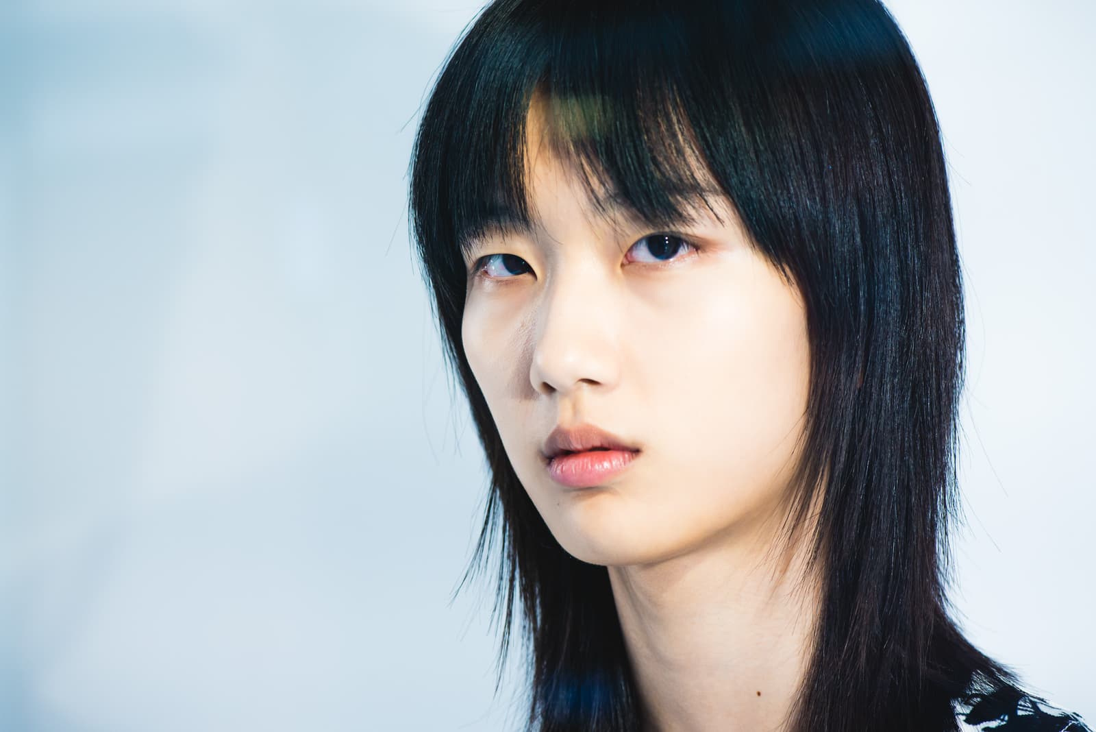 A medium close-up of a person with straight, dark hair and bangs, looking directly into the camera with a neutral expression. The background is a plain, light-grey wall, and the light is soft and even.