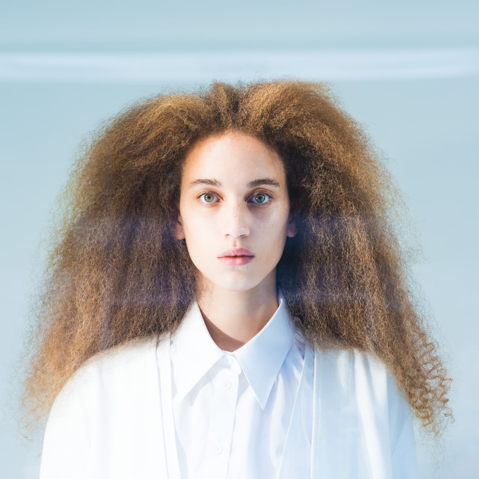 A close-up portrait of a person with voluminous, textured auburn hair against a pale blue background. They are wearing a plain white collared shirt, facing forward with a neutral expression and direct gaze. A soft, horizontal lens flare cuts across the middle of the frame.