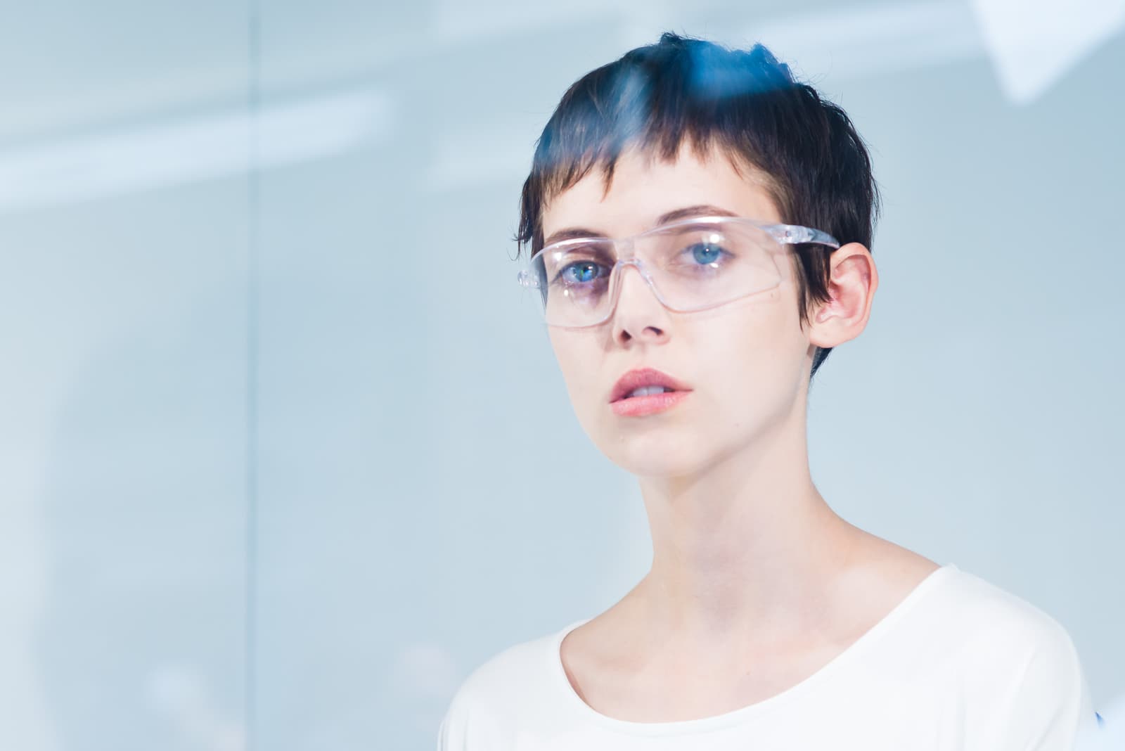 A close-up portrait of a person with short, dark cropped hair, wearing clear plastic safety glasses and a plain white round-neck top. They are positioned behind a reflective, translucent glass surface with light blue lens flares visible at the top. The background is a bright, neutral white wall.