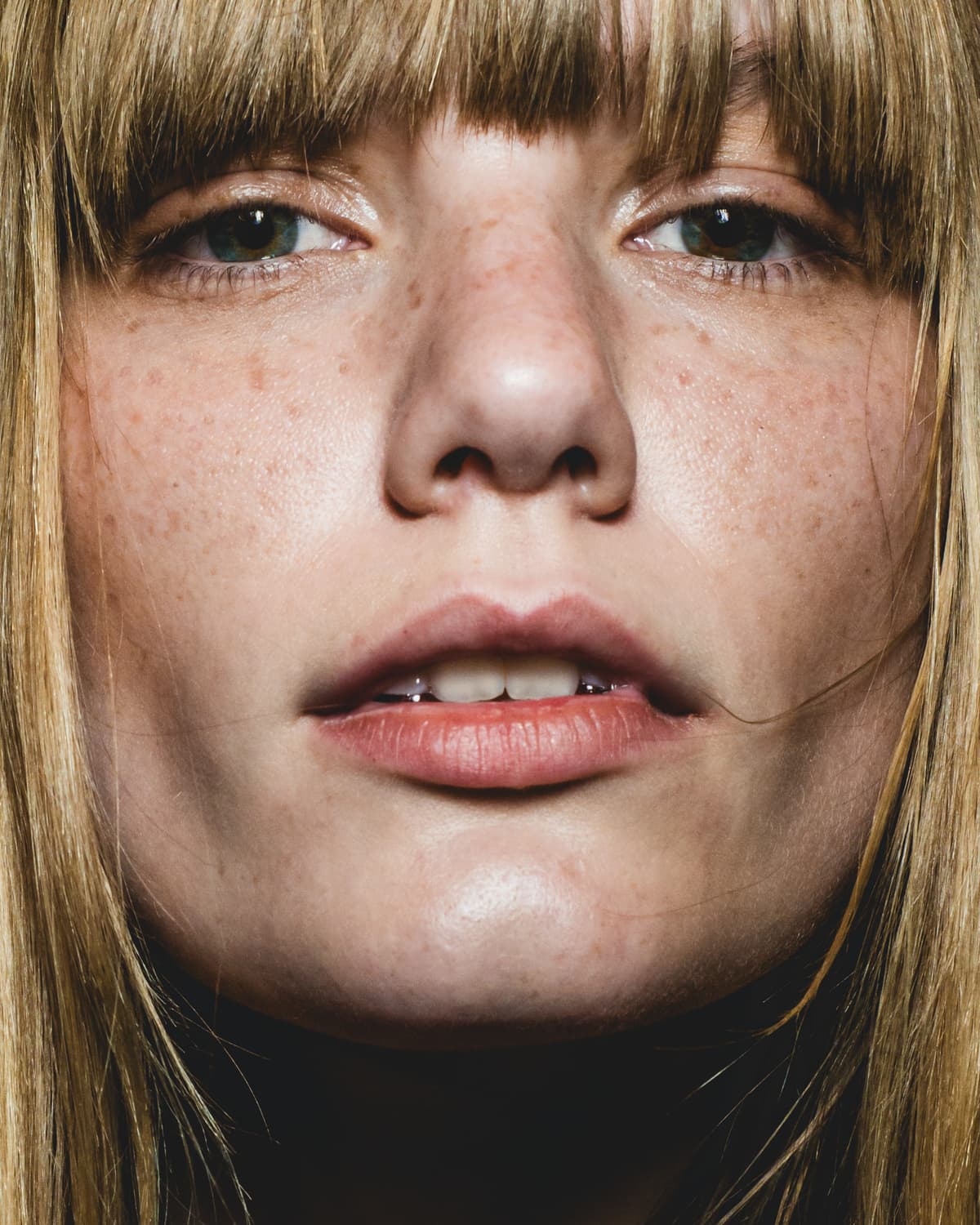 An extreme close-up, frontal portrait of a person with blonde hair, bangs, and noticeable freckles across the bridge of the nose and cheeks. The eyes, with pale irises, gaze directly into the lens. The lips are slightly parted, revealing small dental jewelry.