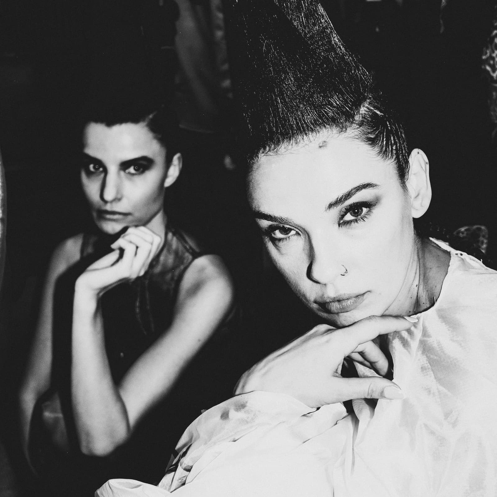 Two people are captured in a close-up, high-contrast black-and-white photograph. The foreground figure, with dark hair styled in a sharp, tall, structured mohawk, looks directly at the camera with a steady gaze and a hand resting under their chin. A second figure is positioned slightly behind and to the left, out of focus, also looking towards the lens. The background is dark and indistinct, suggesting a backstage environment.