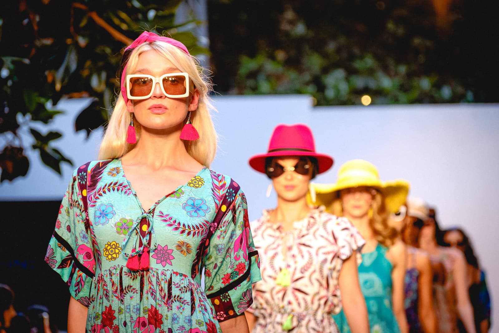 A runway show featuring a line of models walking towards the camera. The model in the foreground wears a bright blue patterned bohemian dress, oversized square-framed sunglasses, and a pink headband with tassel earrings. Behind her, other models follow in a sequence, wearing colourful wide-brimmed hats and vibrant resort-style clothing. The background features a white wall with blurred dark green foliage on the left.
