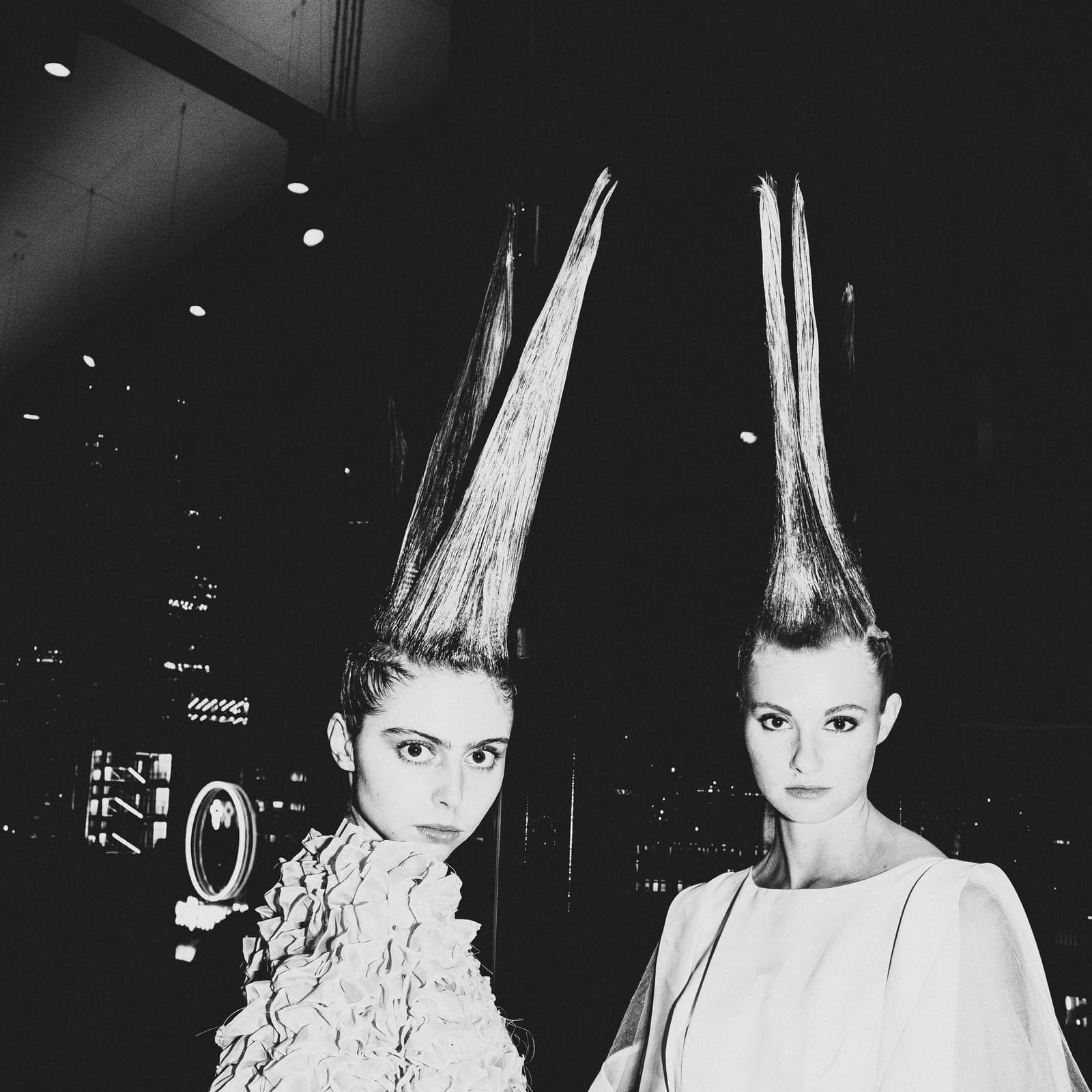 Two people posed side-by-side against a dark background, each sporting extreme, vertically styled hair spikes. The person on the left wears a textured, ruffled collar, and the person on the right wears a smooth garment with thin piping detail. Both gaze directly at the camera in a dimly lit, possibly indoor industrial or backstage environment.