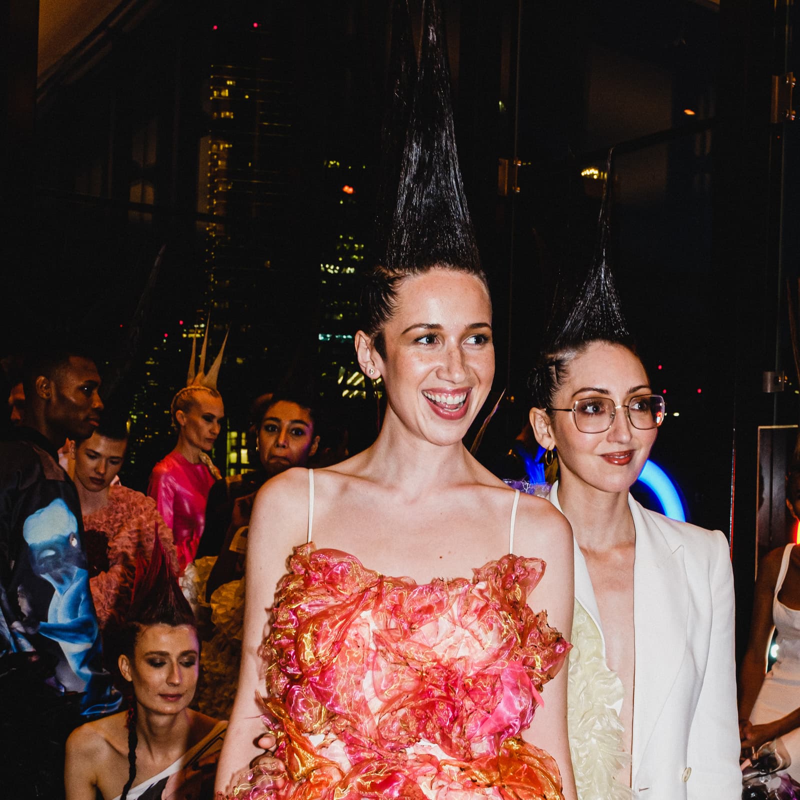 A medium shot taken backstage at a fashion show, showing two people in the foreground. The person on the left wears a dress with layers of ruffled, pink and gold iridescent fabric and has hair styled into a high, stiff vertical point. The person to the right wears a white blazer, gold-rimmed glasses, and has hair similarly styled into a high, stiff vertical point. Behind them are several other individuals in the dimly lit backstage area, some with similar gravity-defying hairstyles and vibrant clothing.