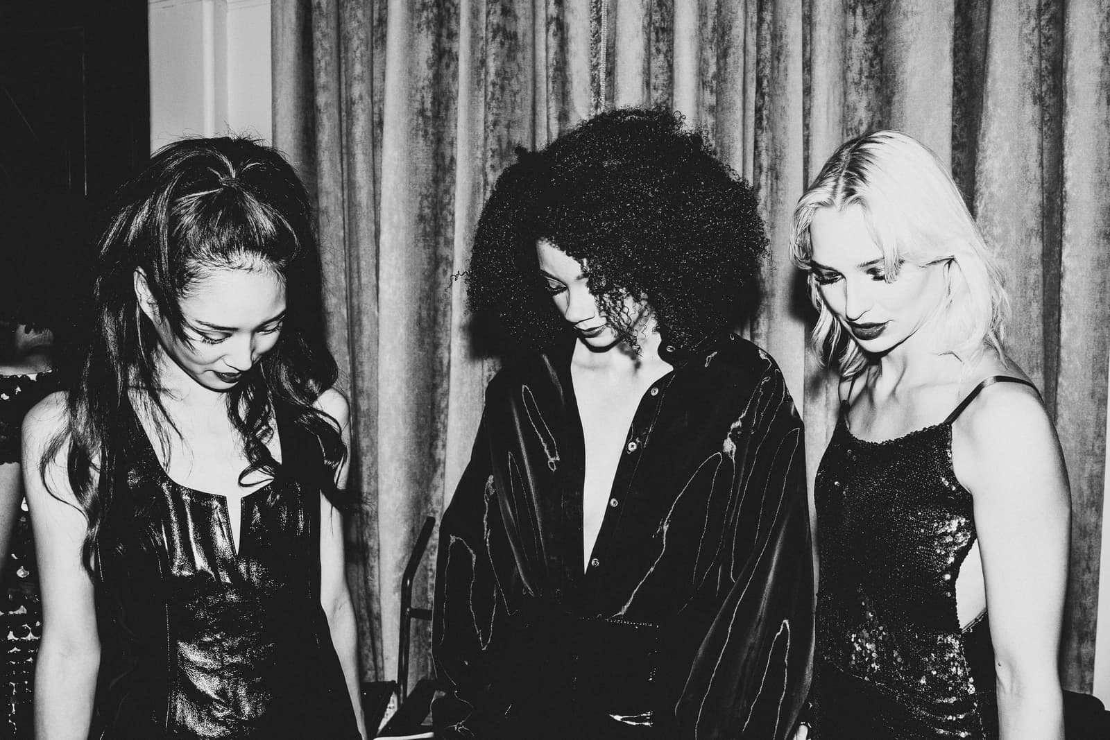 Three individuals standing in a row, captured in a black and white medium shot. They are all looking downward with neutral expressions. The individual on the left has long, dark hair tied up, wearing a dark, sleeveless, textured top with a neckline detail. The individual in the center has dark, voluminous curly hair and wears a dark, button-down collared shirt with thin, light-colored stitched lines. The individual on the right has shoulder-length, lighter hair and is wearing a dark, sequined camisole top. The background is a simple, hanging fabric curtain with vertical folds.