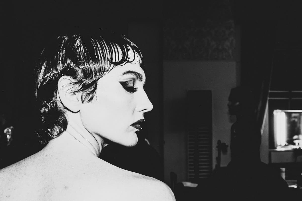 A black-and-white profile view of a person with dark, short hair and bold, dark eye makeup, looking away from the camera. The subject is bare-shouldered, captured in a dimly lit indoor setting with a blurred background.