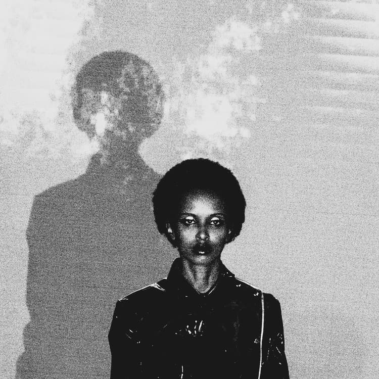 A black-and-white medium shot of a person with dark hair, wearing a shiny dark jacket, looking directly at the camera. A large, out-of-focus shadow of the same person is projected onto the light-colored wall behind them.