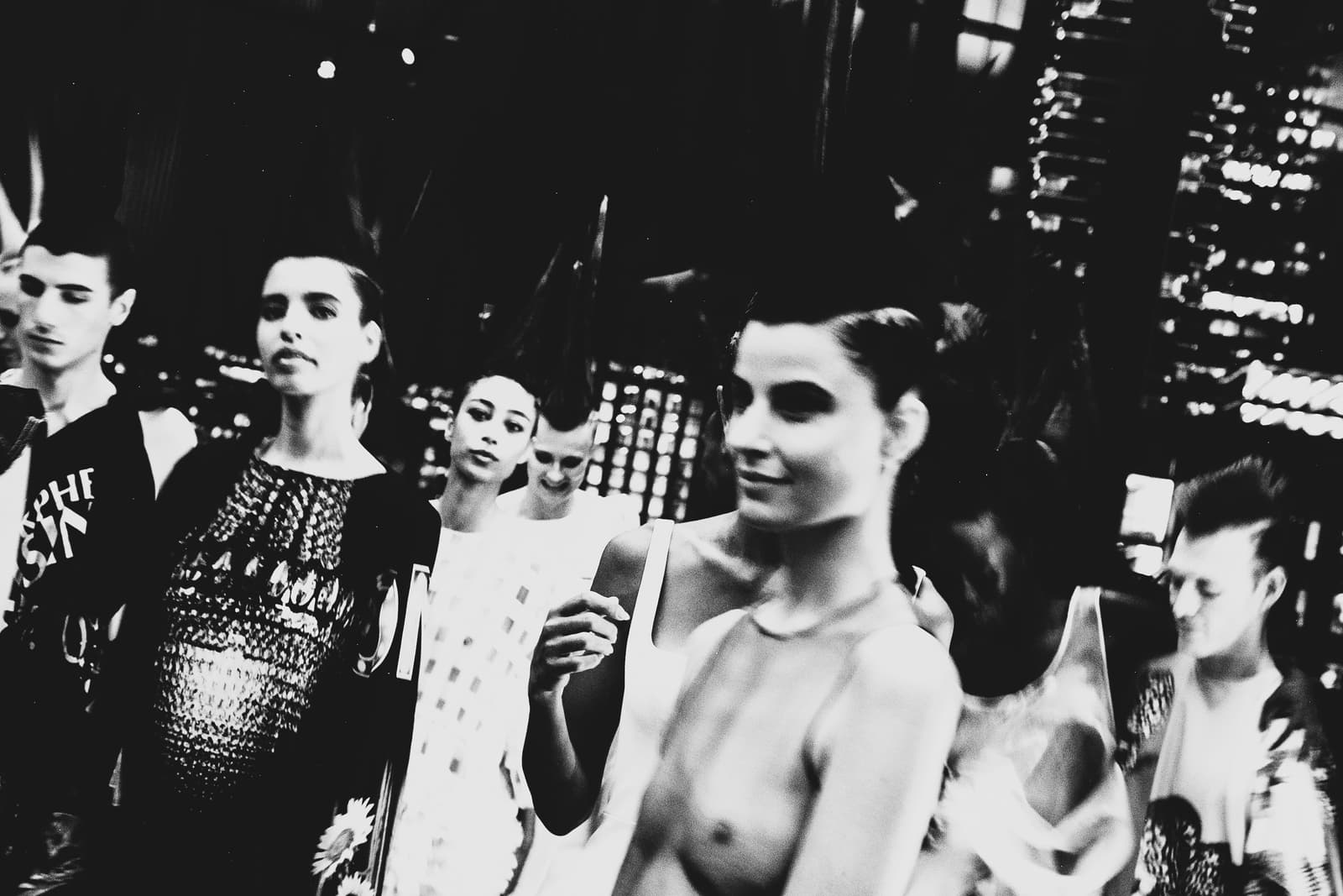 A black-and-white medium shot of several models walking in a backstage area during a fashion show, with one model in the immediate foreground partially out of focus, wearing a light-coloured garment, and others in the background with varied expressions and styled hair.