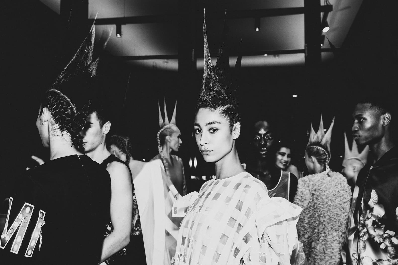 A black-and-white backstage scene at a fashion show featuring several models with elaborate, tall spiked hairstyles. In the center, a person in a white, grid-patterned garment with oversized sleeves looks directly at the camera. The setting is dimly lit with industrial overhead lighting, and other figures are partially visible in the crowded background.