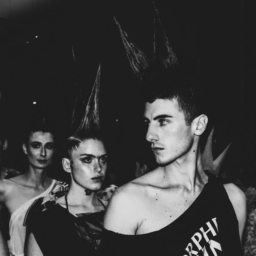 A black-and-white, medium close-up shot from backstage at a fashion show. Three people are positioned in the frame, wearing dark sleeveless garments and dramatic, spiked, vertical hairstyles. The person in the foreground is in profile, looking toward the center, while the person behind them looks directly at the camera with dark eye makeup. The setting is dimly lit, with the focus on the hair texture and the stark contrast of the black clothing against their skin.
