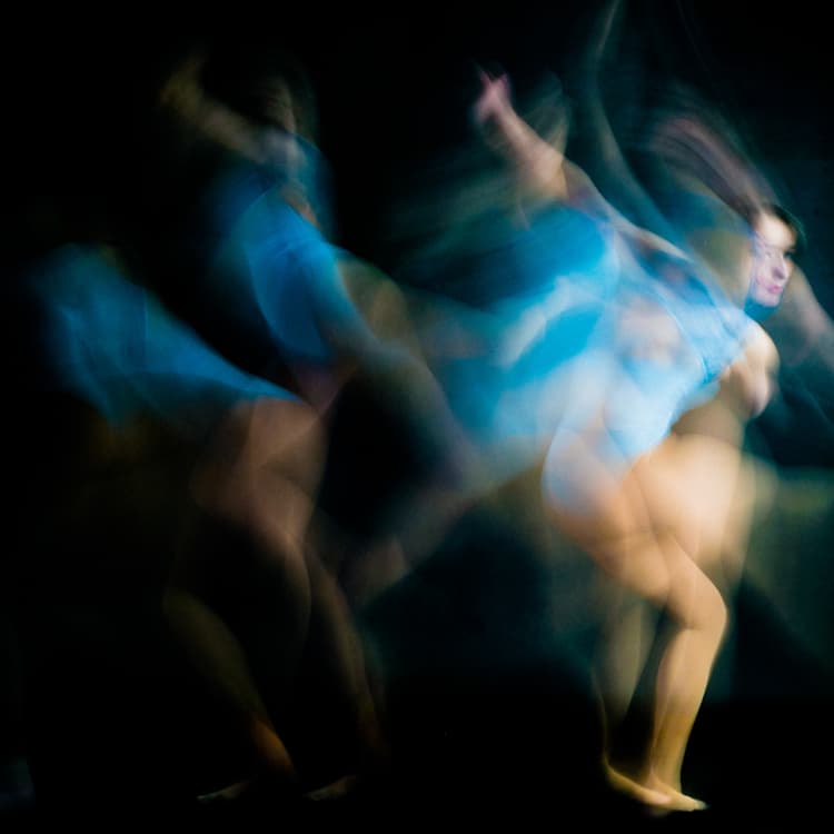 A long-exposure photograph of a person in a blue leotard moving across a dark stage, leaving multiple blurred motion trails behind their body, with one sharp frame of their face visible on the right side.