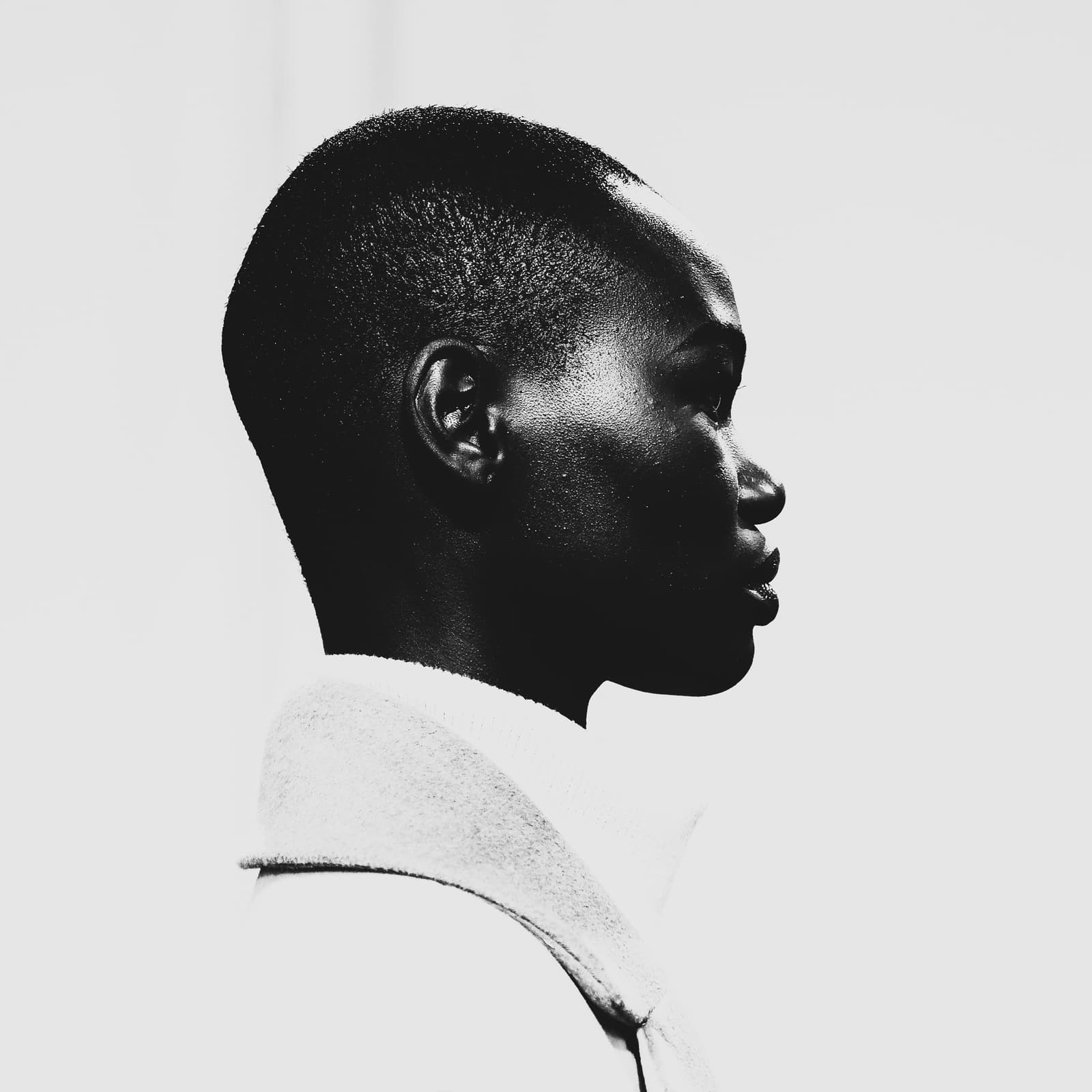 A high-contrast black and white profile portrait of a person with a shaved head, facing right. Strong light creates deep shadows on the face, emphasizing the jawline and the texture of the skin. The person wears a plain high-collared garment against a bright, featureless background.